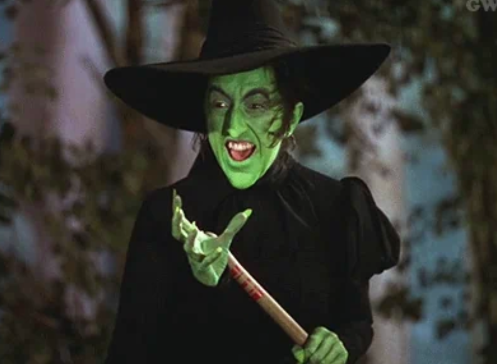 A person in a witch costume with green makeup, a pointed hat, and holding a broomstick, laughing dramatically in a cinematic setting
