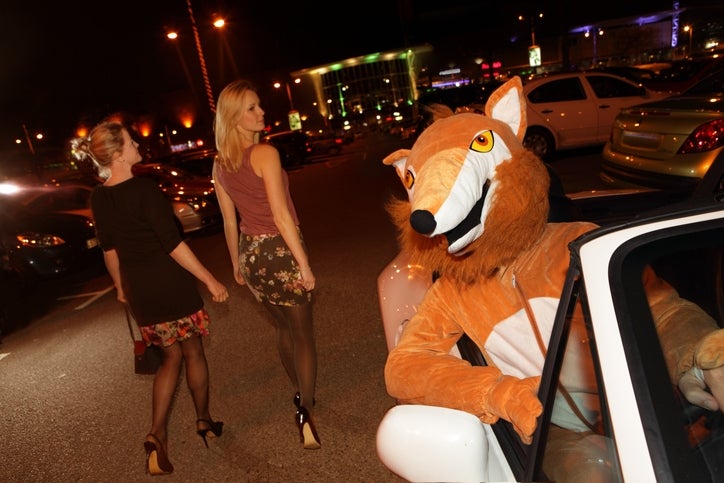 Person in a fox costume sits in a car while two women walk by in an evening parking lot, smiling and dressed in stylish casual outfits
