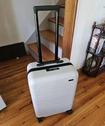 A sleek, modern suitcase with wheels and a telescopic handle, positioned in a cozy home setting near a staircase and bookshelf