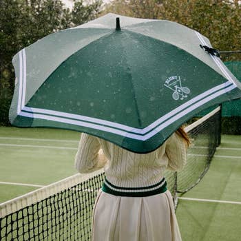 Person holding a tennis-themed umbrella on a rainy court, wearing a cable knit sweater and skirt