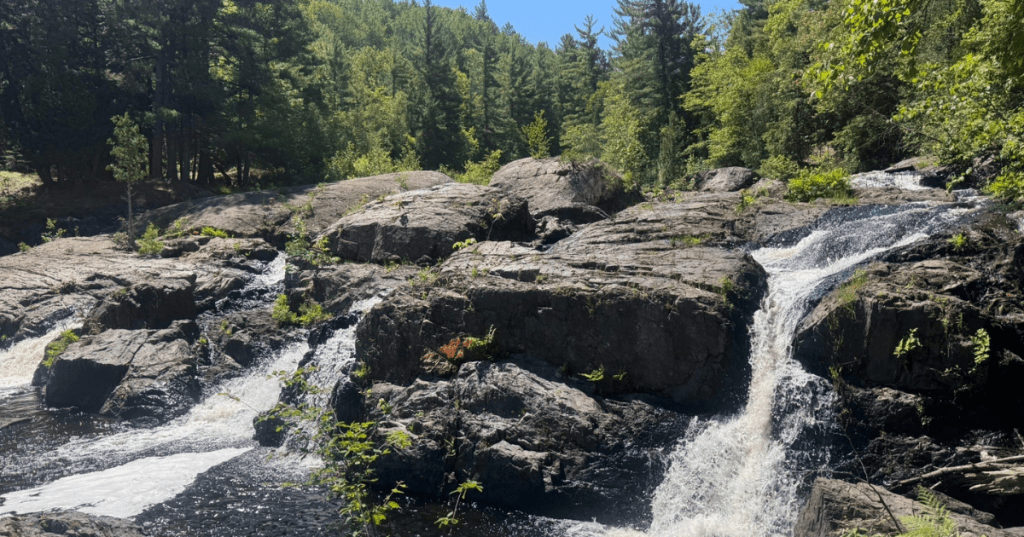 Split waterfall rushing over dark, rocky terrain surrounded by a thick summer forest in Marquette, MI.