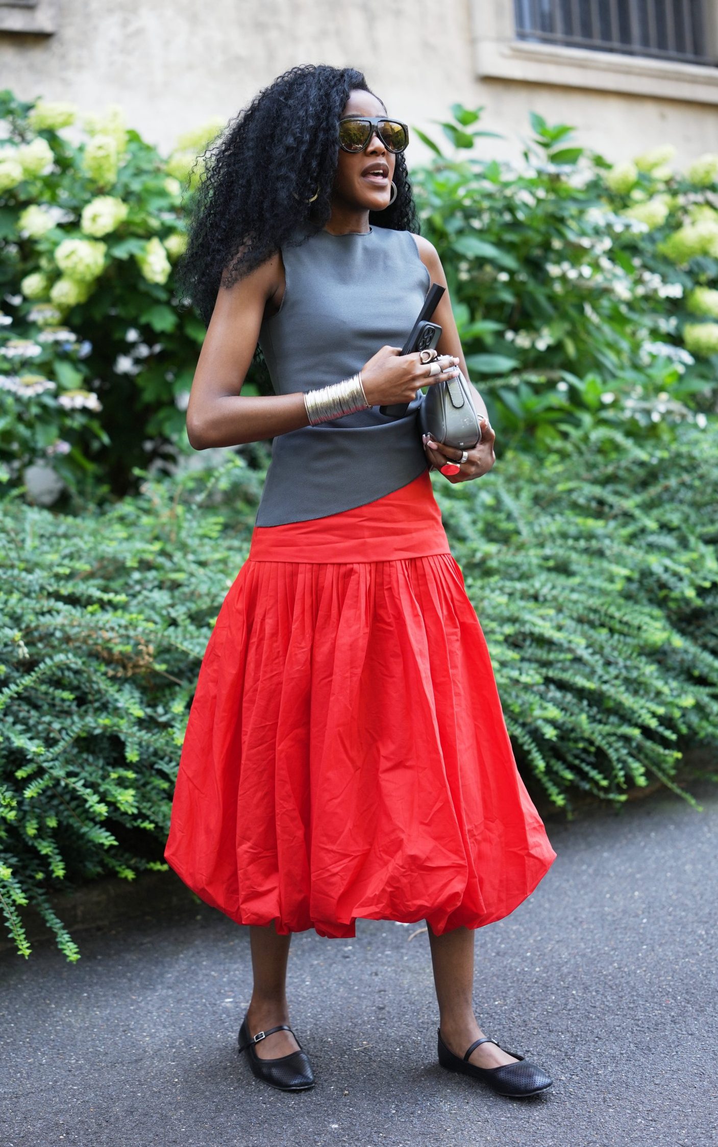A guest wears long curly black hair, oversized black square sunglasses, a sleeveless gray asymmetric top, a silver large cuff bracelet, a neon red pleated maxi skirt, a silver varnished leather crossbody bag, black Mary Jane flats shoes, outside Taakk, during Paris Fashion Week in June 2026