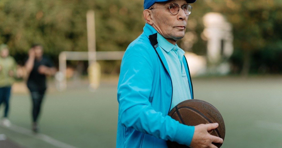 Elderly man with glasses and cap holding a basketball on an outdoor court.