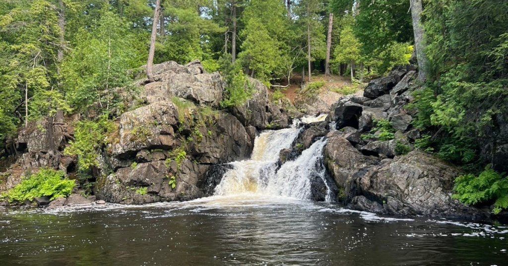 Powerful rushing waterfall framed by rugged rock formations and dense green forest in Marquette, Michigan.