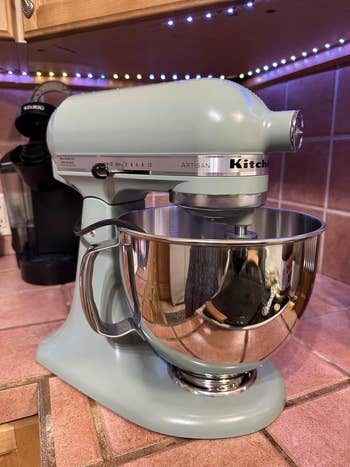 Stand mixer on kitchen counter with shiny stainless steel bowl, next to a coffee maker and under colorful LED lights