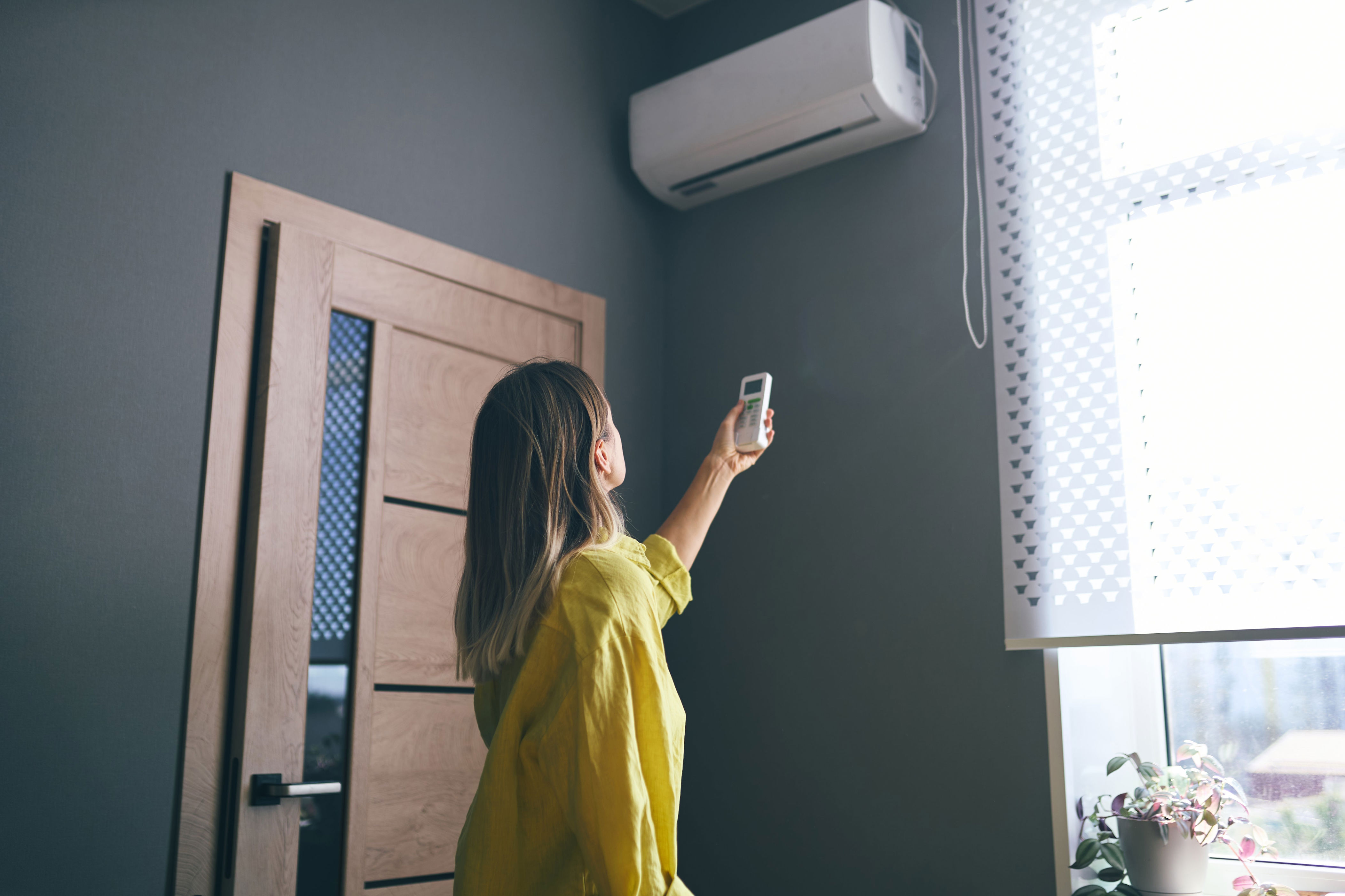A person in casual clothes adjusts an air conditioner with a remote in a room with a door and window blinds