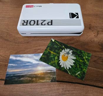 Portable photo printer on a wooden table with two printed photos: a scenic sunrise and a close-up of a daisy