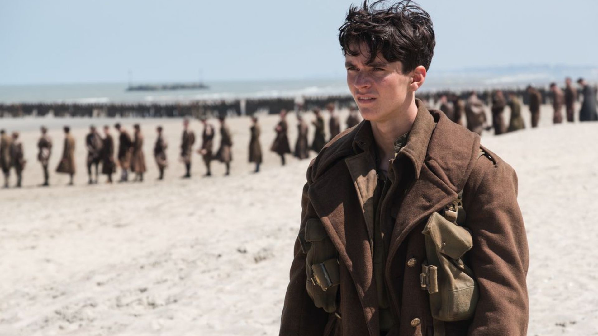 Fionn Whitehead as Tommy standing on the beach with soldiers behind him in a scene of Christopher Nolan's movie, Dunkirk