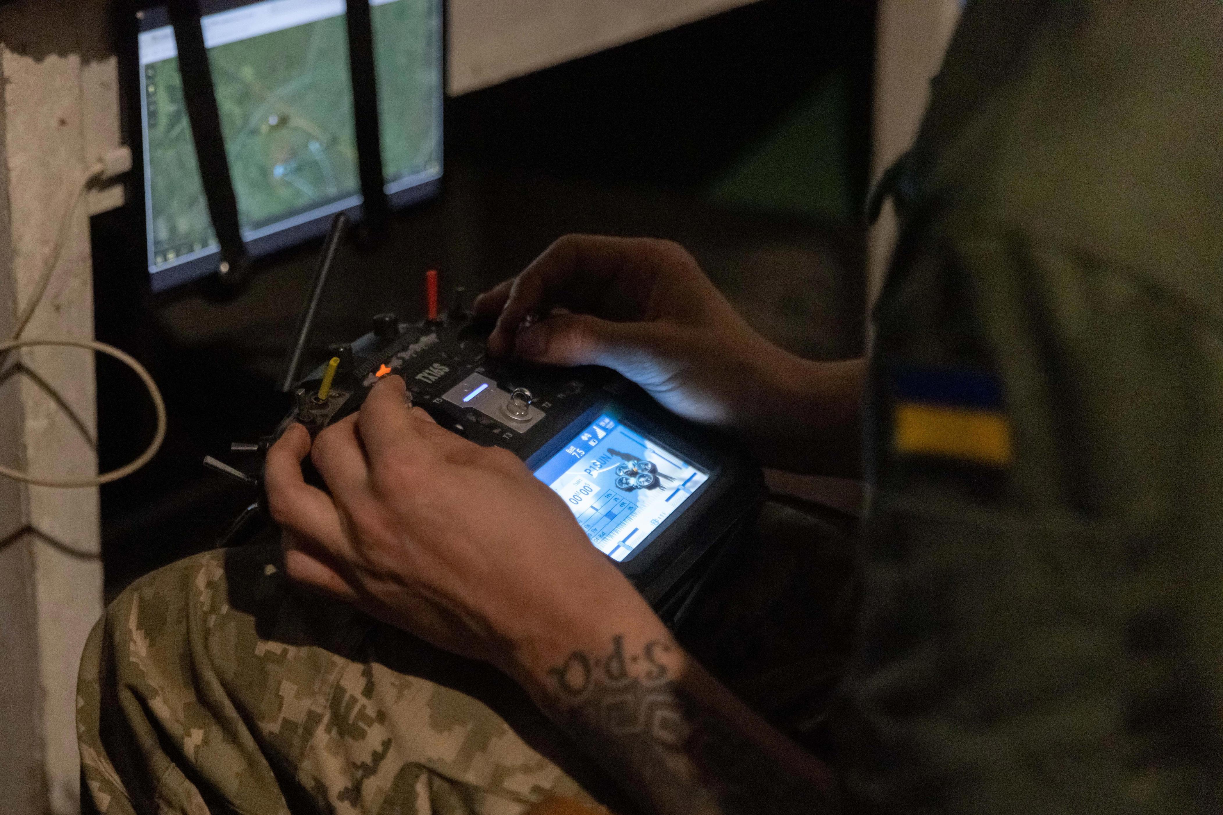 Uniformed person with a Ukrainian flag patch operates a drone controller beside monitors showing map and video feeds.