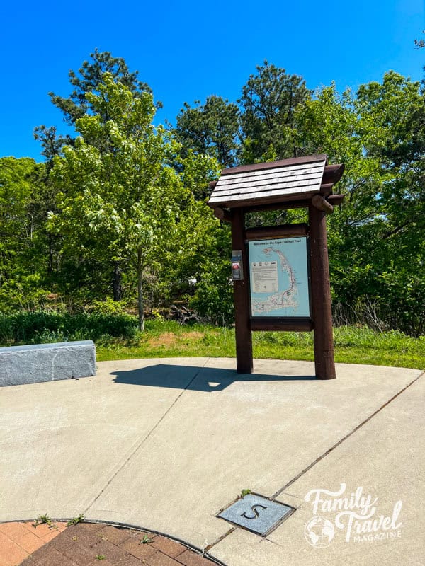 A sign for the Cape Cod rail trail sits on a rounded concrete area among trees at the entrance of the Cape Cod rail trail.
