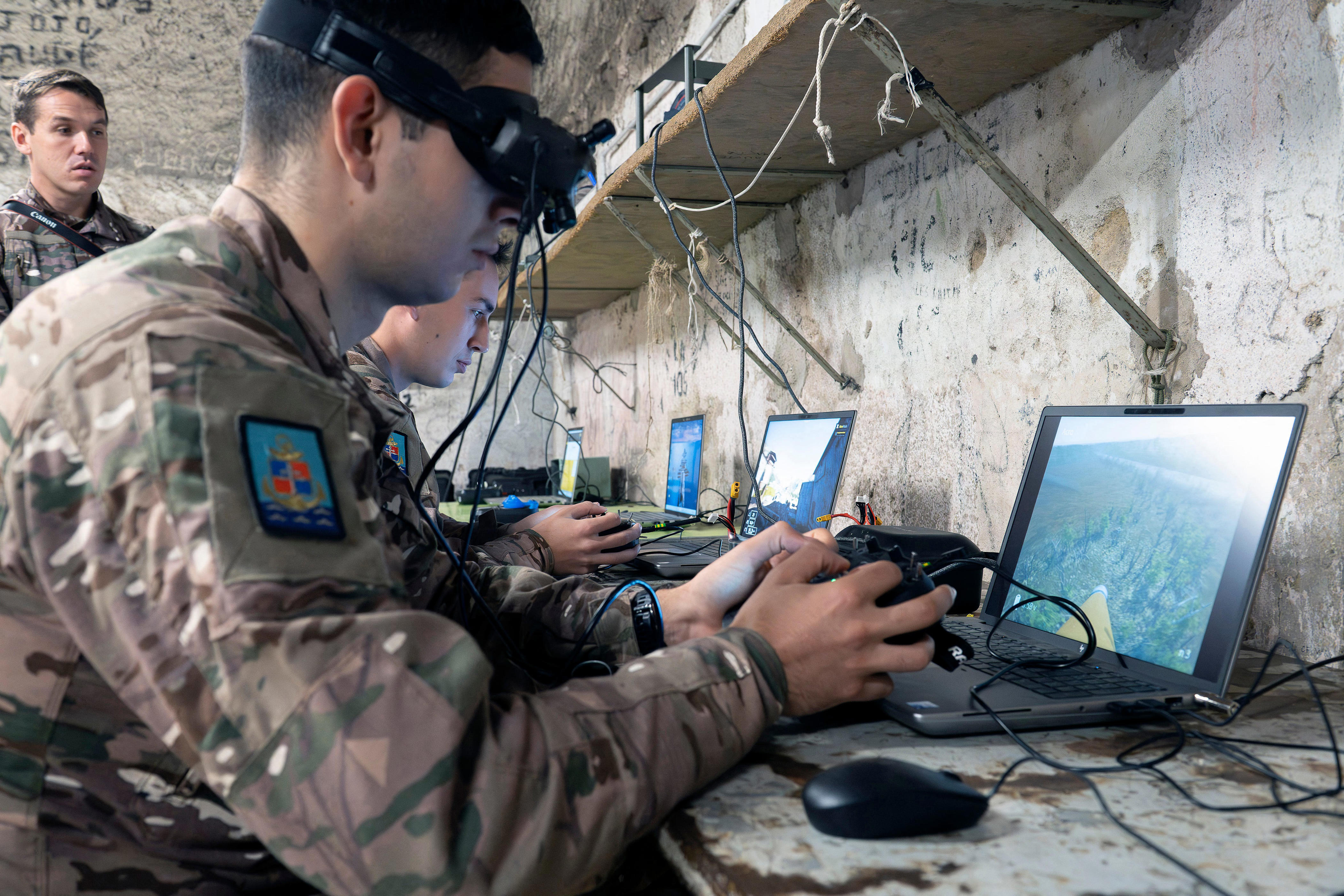 Uniformed personnel operate laptops and controller equipment at a makeshift workstation beside a concrete wall.