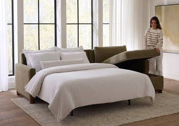 Person lifting the storage lid of a stylish sofa bed in a modern, minimalist bedroom setting with large windows and a framed abstract art piece