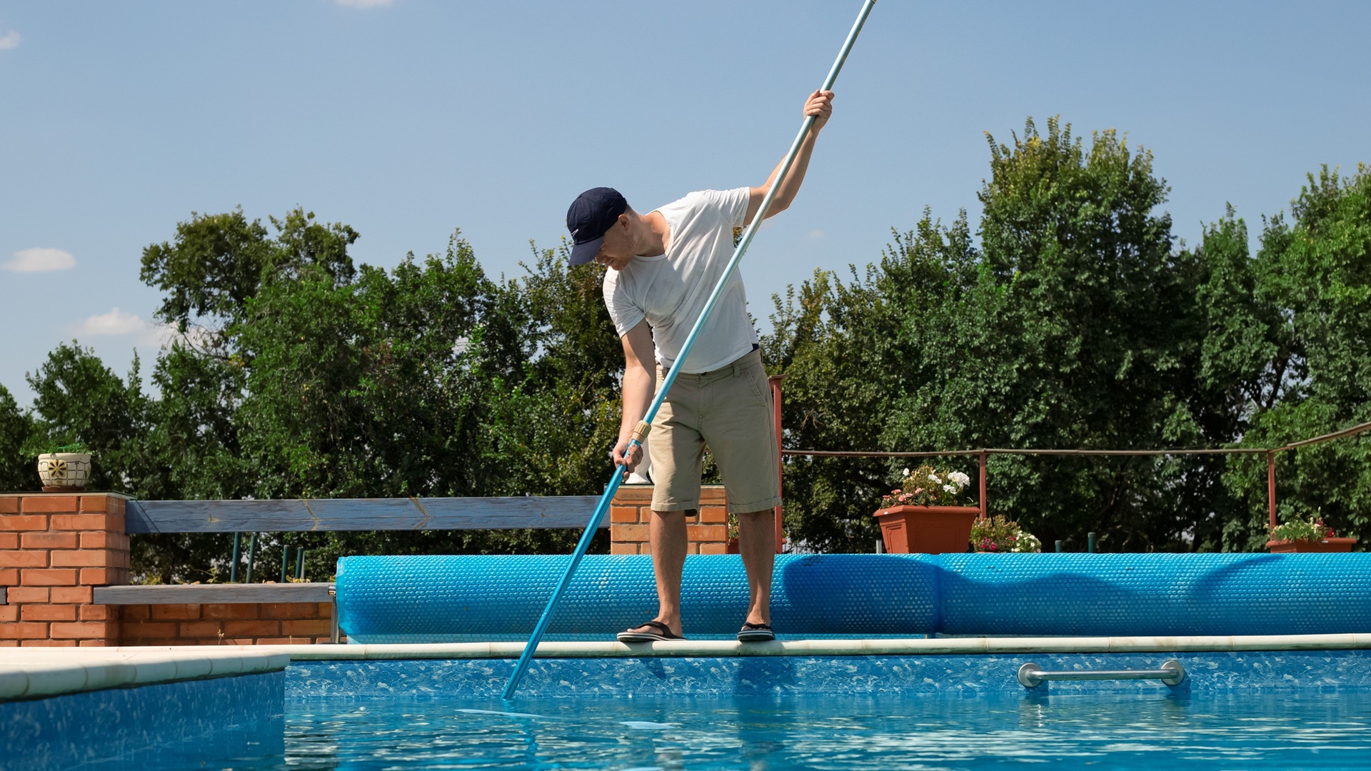 Discover a man in a baseball cap and T-shirt cleans a pool using a large pole with a net attached