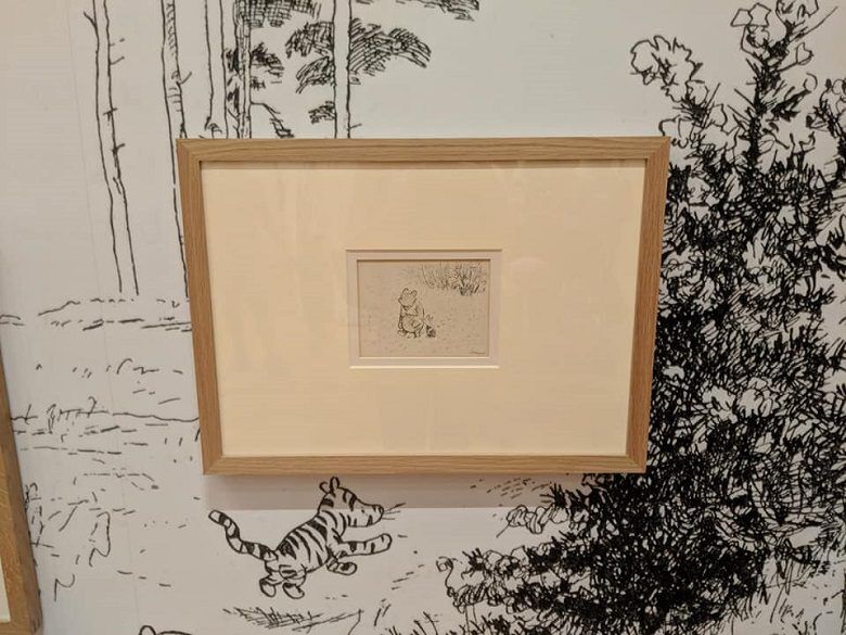 A small framed sketch hangs against wallpaper covered with black line drawings of trees and familiar storybook characters from Winnie-the-Pooh including a bouncing tiger. The delicate artwork is centered in a wide cream mat and simple wood frame at the High Museum of Art in Atlanta, Georgia.