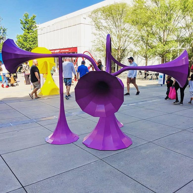 Sonic Playground exhibit in Sifly Plaza at the High Museum of Art in Atlanta, Georgia. A bright purple outdoor sculpture shaped like several oversized trumpet horns stands in a museum courtyard. Visitors walk around the installation, with a yellow sculpture, leafy trees, and a white building in the background.