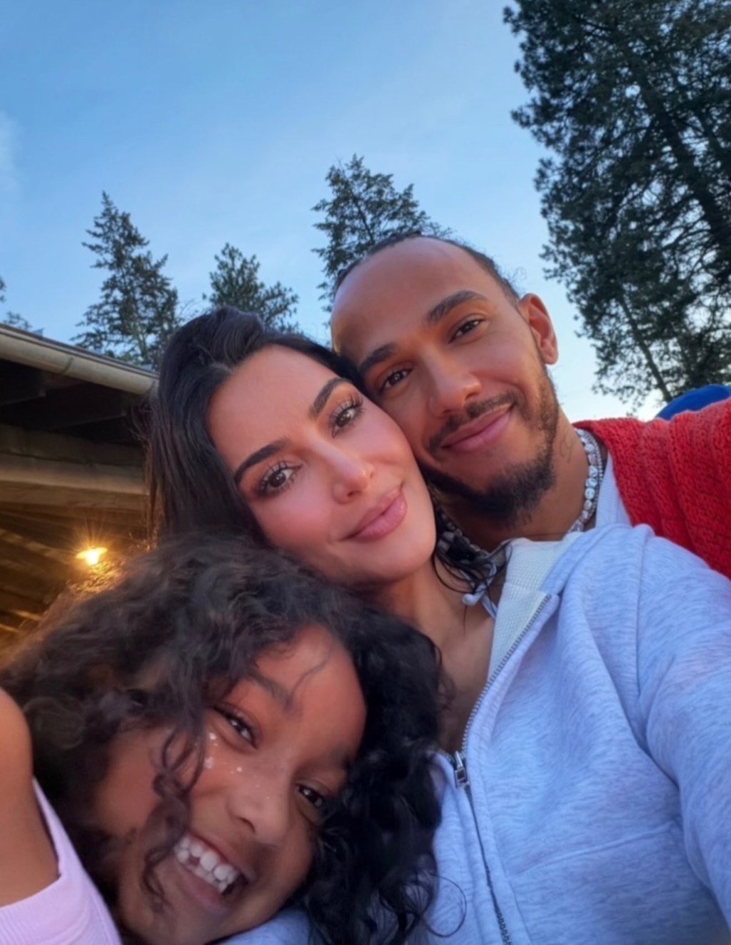 Kim Kardashian, Lewis Hamilton, and a child smiling for a selfie outdoors.
