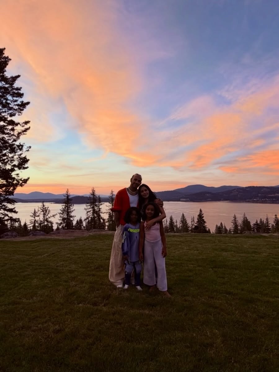Kim Kardashian and Lewis Hamilton with two children, posing for a photo in front of a scenic sunset over a lake.