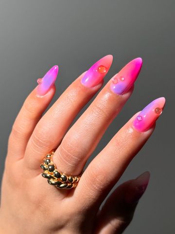 Well manicured hand with gold rings glowing nail art design with pink and purple hues and glossy 3D accent details
