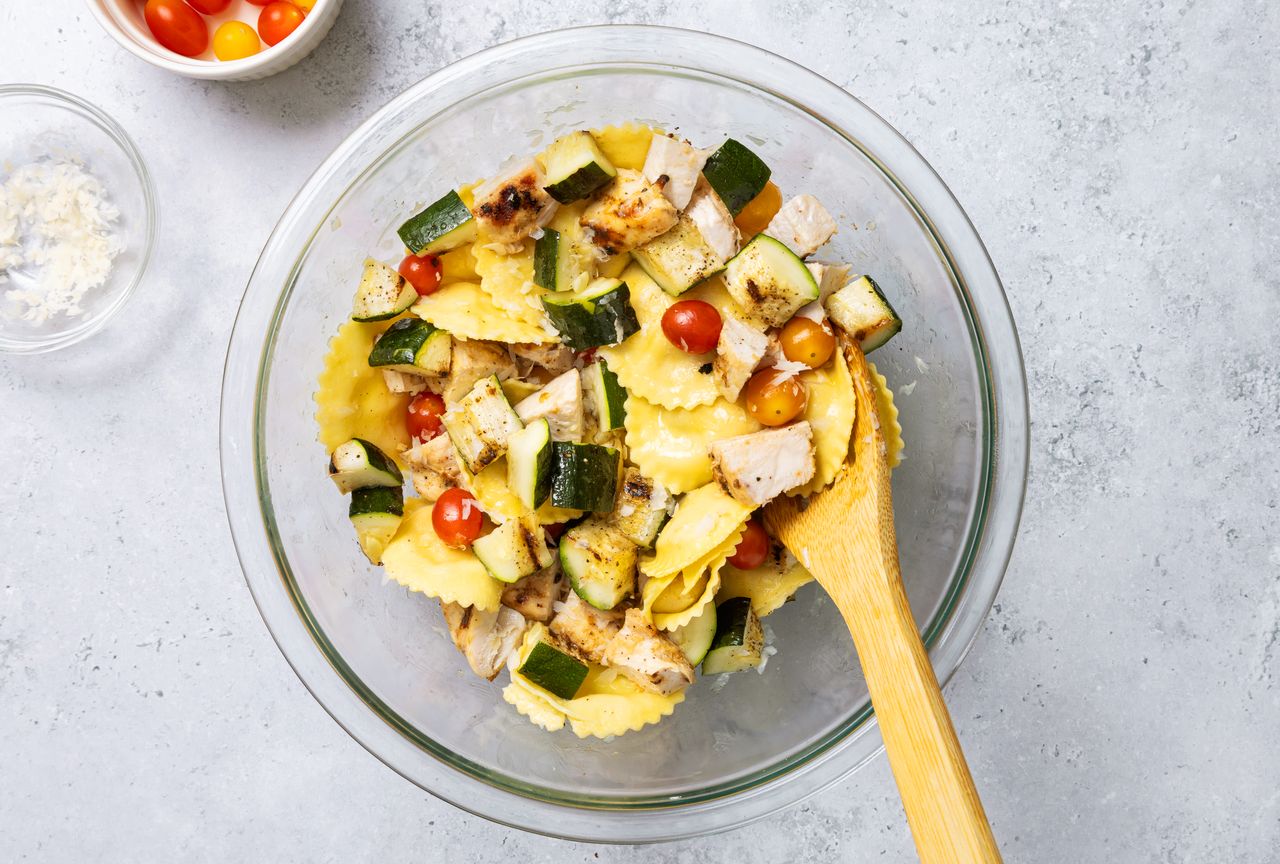 A glass bowl containing a pasta salad with ravioli grilled vegetables and cherry tomatoes a wooden spoon in the bowl