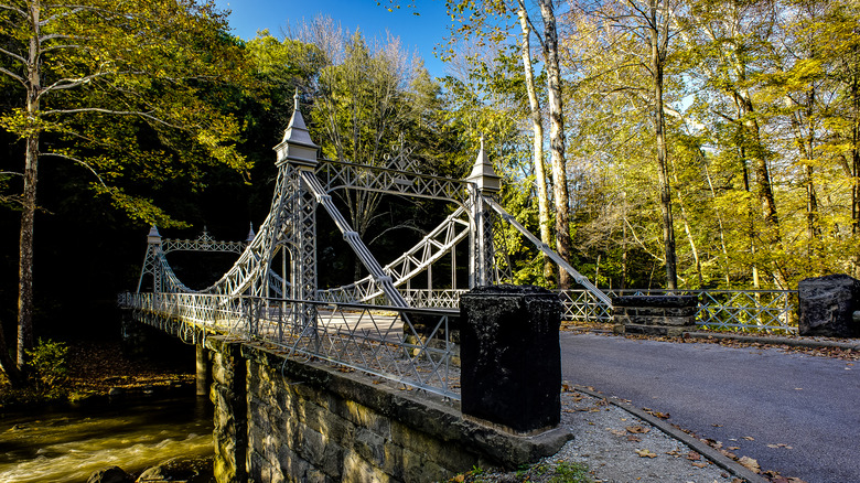 A suspension Bridge in Mill Creek Park, a white-painted metal structure with a gravel path running across it over a flowing creek, with trees surrounding it under a blue sky