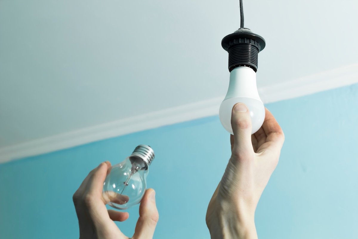 Person holds incandescent light bulb in one hand, replacing it wtih an LED light bulb in an overhead socket.