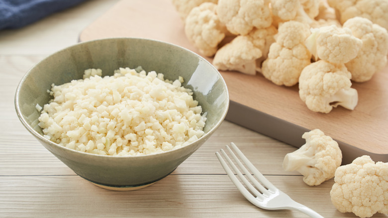 Cauliflower rice in bowl next to cauliflower on cutting board