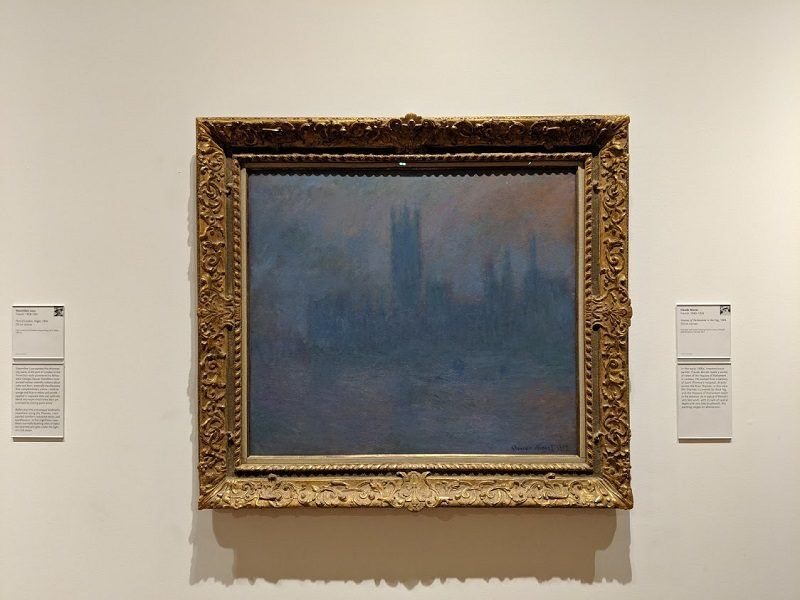Houses of Parliament in the Fog, by Claude Monet, at the High Museum of Art in Atlanta, Georgia. A hazy blue gray painting of a city skyline and water hangs in an ornate gold frame on a museum wall. Small information labels are mounted on both sides of the artwork.