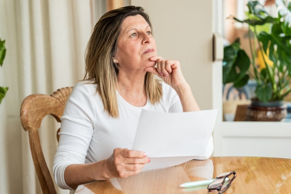 A thoughtful woman sits at a table holding a document, hand on chin, looking upwards.