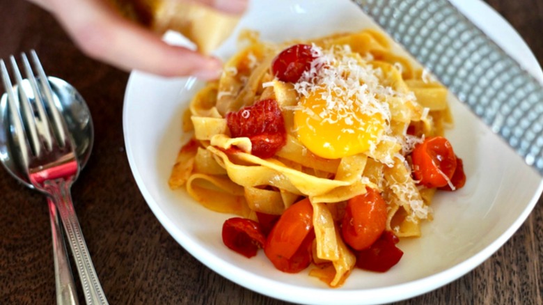 Plate of pasta with tomatoes and egg yolk, with a hand adding parmesan cheese