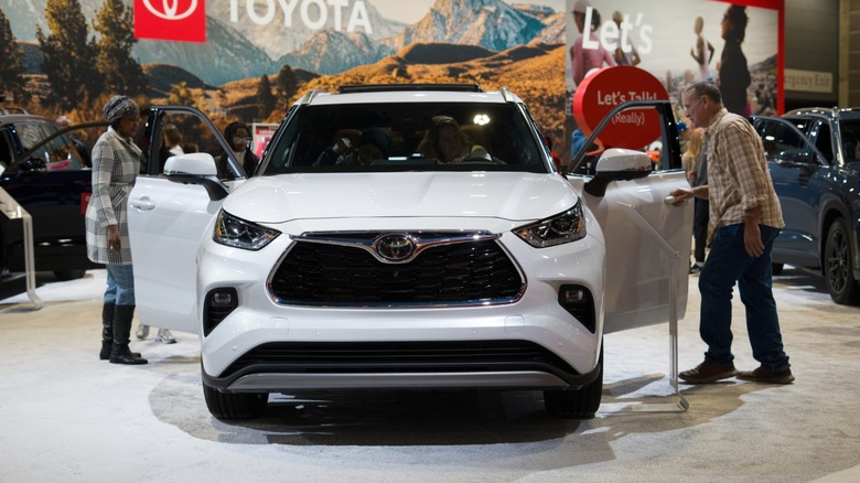 Front view of a white Toyota Highlander displayed at an auto show, with both front doors open as attendees examine the SUV