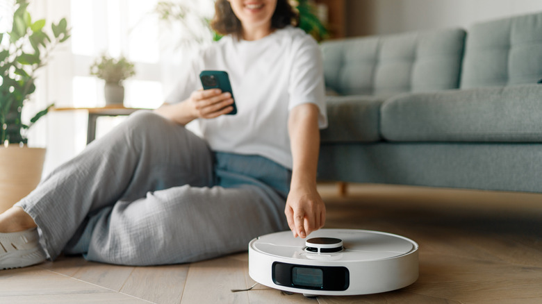 A woman sitting on the floor smiling while holding her phone, a robot vacuum in front of her.