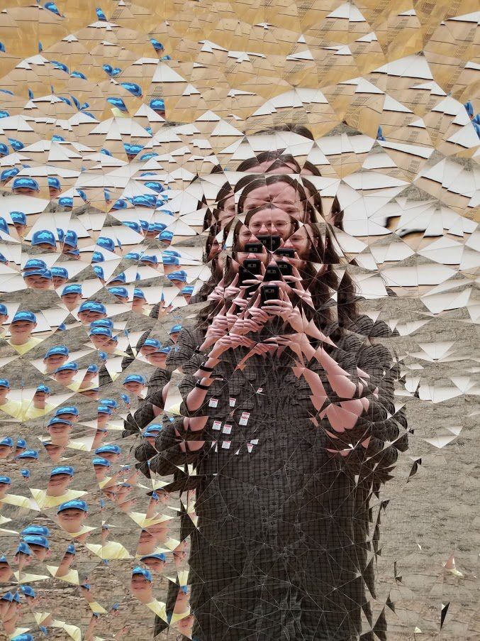 Concave Dish Sculpture at the High Museum of Art in Atlanta, Georgia. A fragmented mirror installation multiplies the reflection of a person holding a phone, while another visitor in a blue cap appears repeatedly across the left side. Hundreds of angular mirrored pieces create a kaleidoscope effect against a tan and white background.