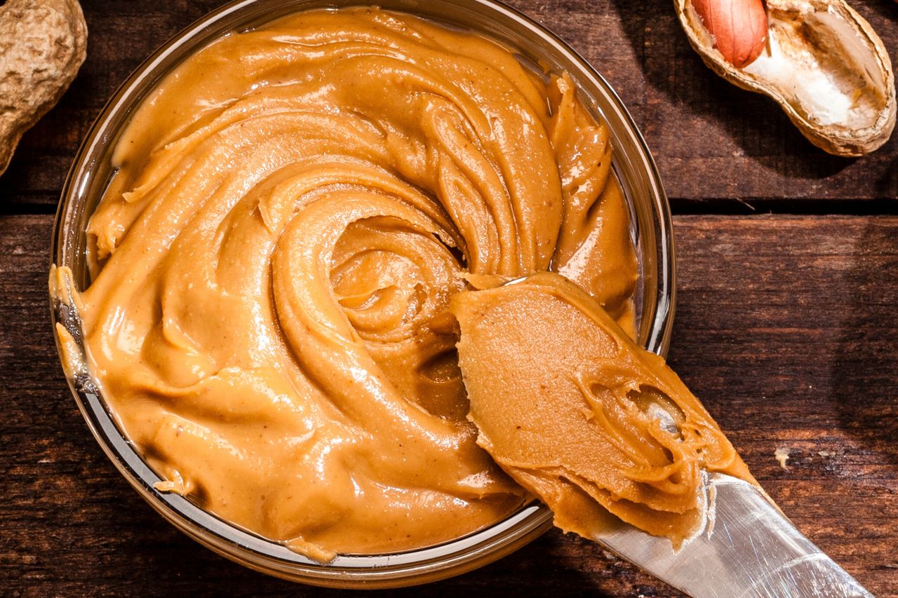 A closeup of peanut butter in a bowl with a butter knife spreading a portion some peanuts visible in the background