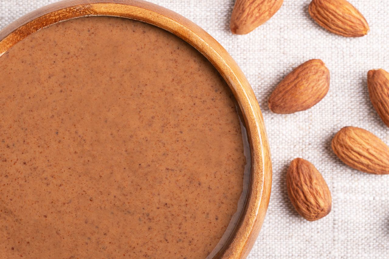 Wooden bowl of almond butter with almonds scattered nearby on a light surface