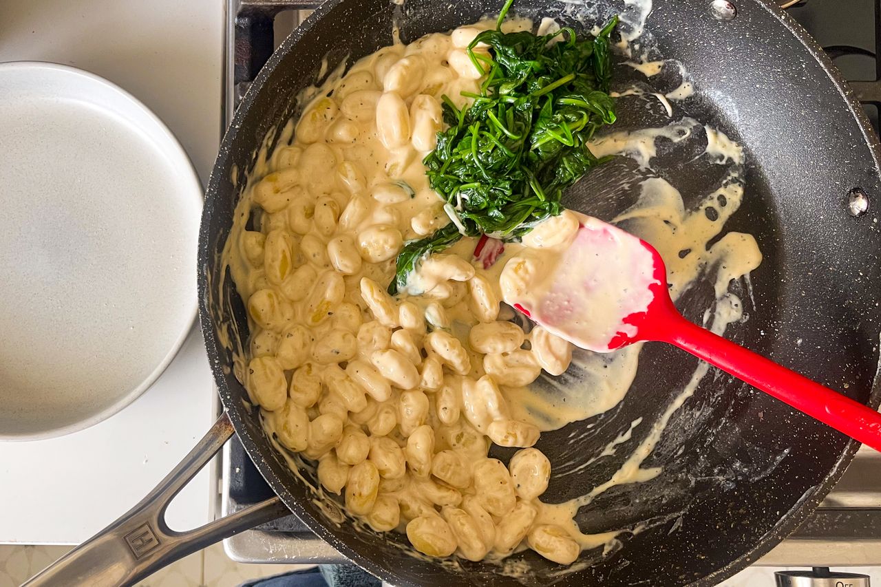 A skillet of cooked gnocchi with cream sauce and spinach being stirred with a red spatula