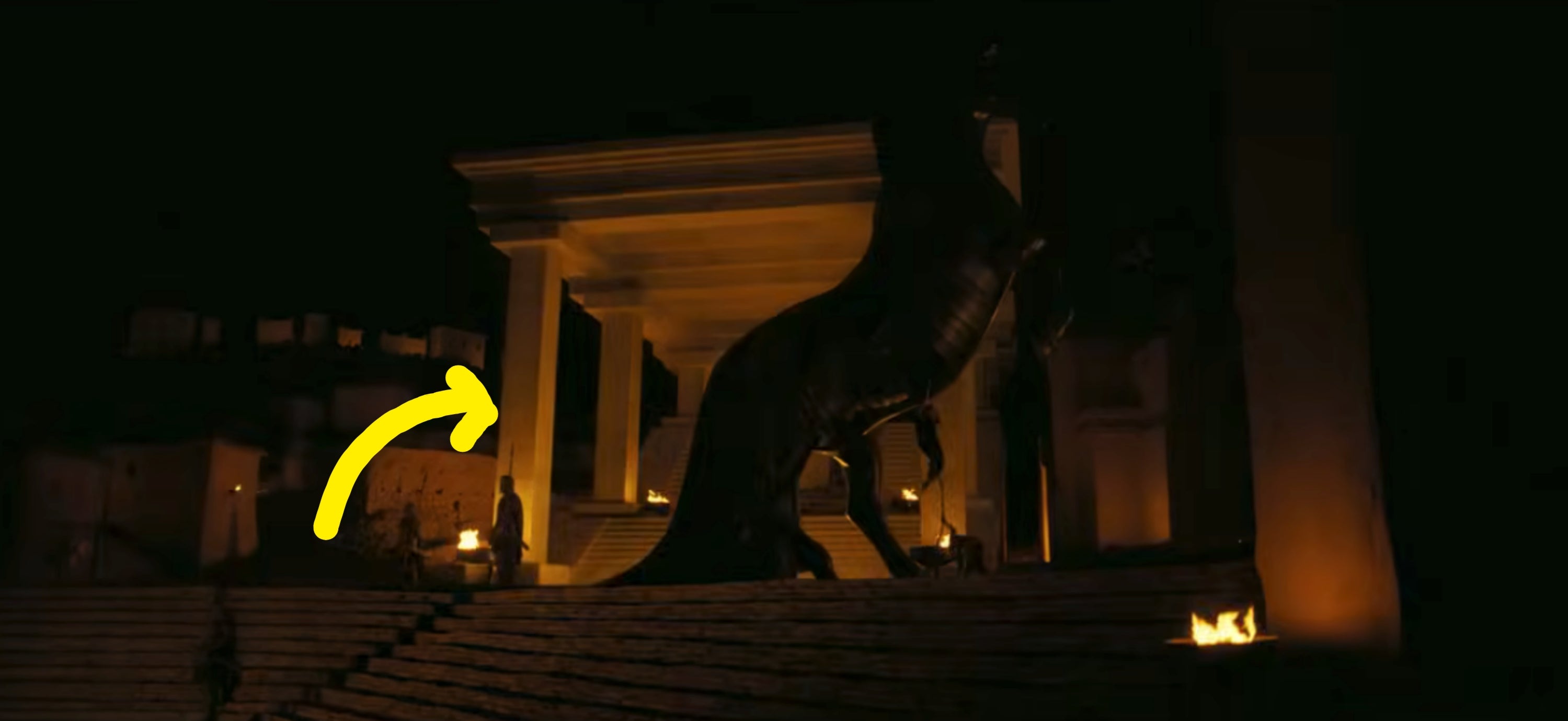 A large Trojan horse stands on stone steps at night, surrounded by torches in front of a classical building