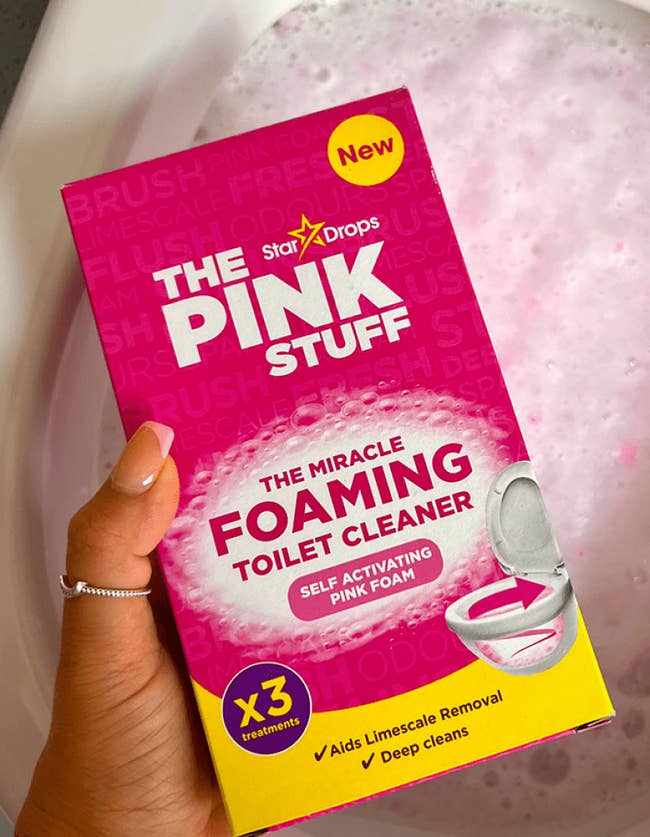 Hand holding The Pink Stuff foaming toilet cleaner box over a bubbly toilet. Text highlights its features: limescale removal and deep cleaning