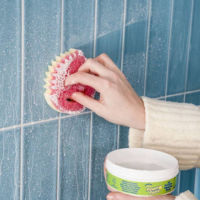 Person cleaning blue tiled wall with a scrub sponge and tub of Power Paste