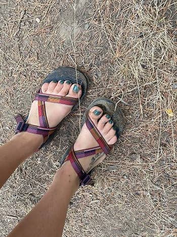 Person wearing strappy, casual sandals with a tattoo visible on their foot stands on dry grass and dirt