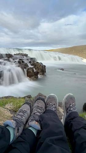 Two people wearing hiking boots sit in front of a scenic waterfall, which cascades over rocks into a gentle river