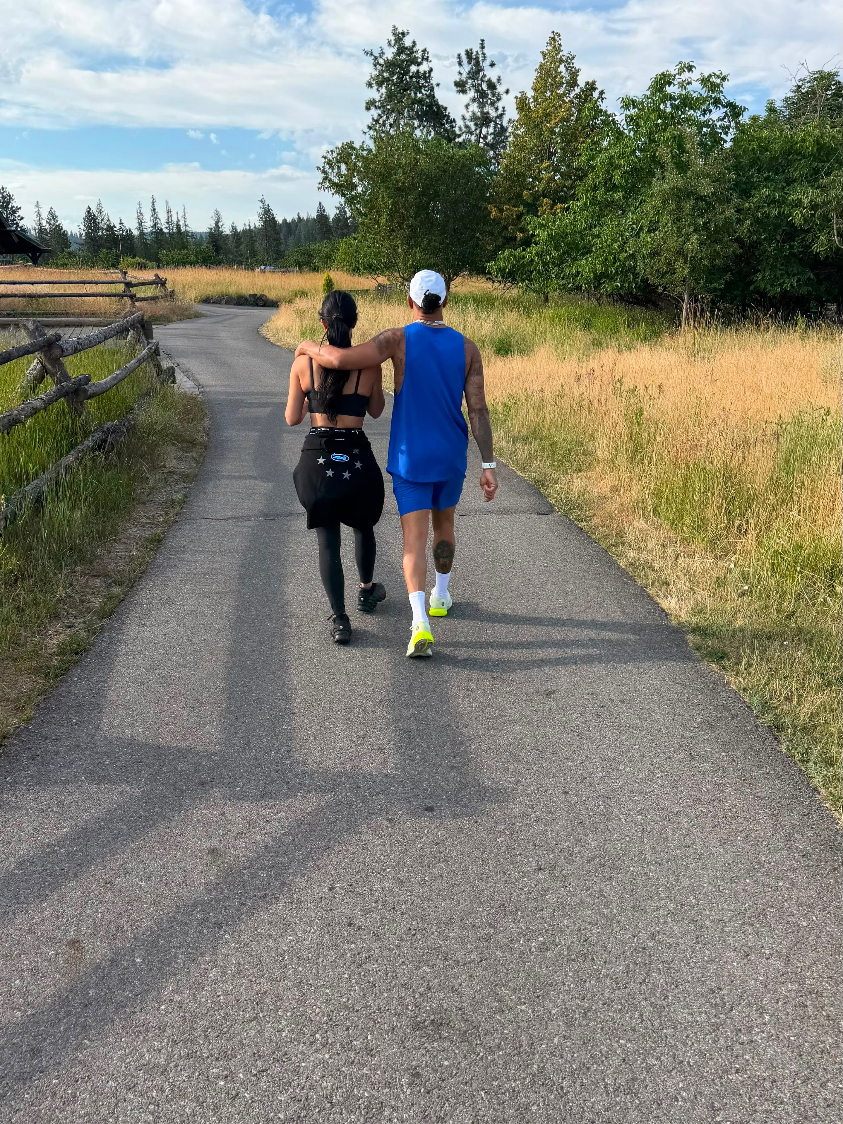 Kim Kardashian and Lewis Hamilton walking arm-in-arm down a path, with a wooden fence and grassy fields on either side.