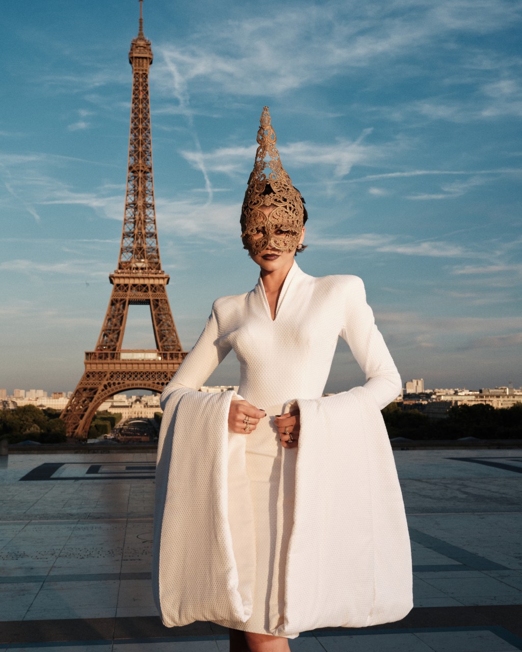 Zendaya poses in a white dress and golden mask in front of the Eiffel Tower.