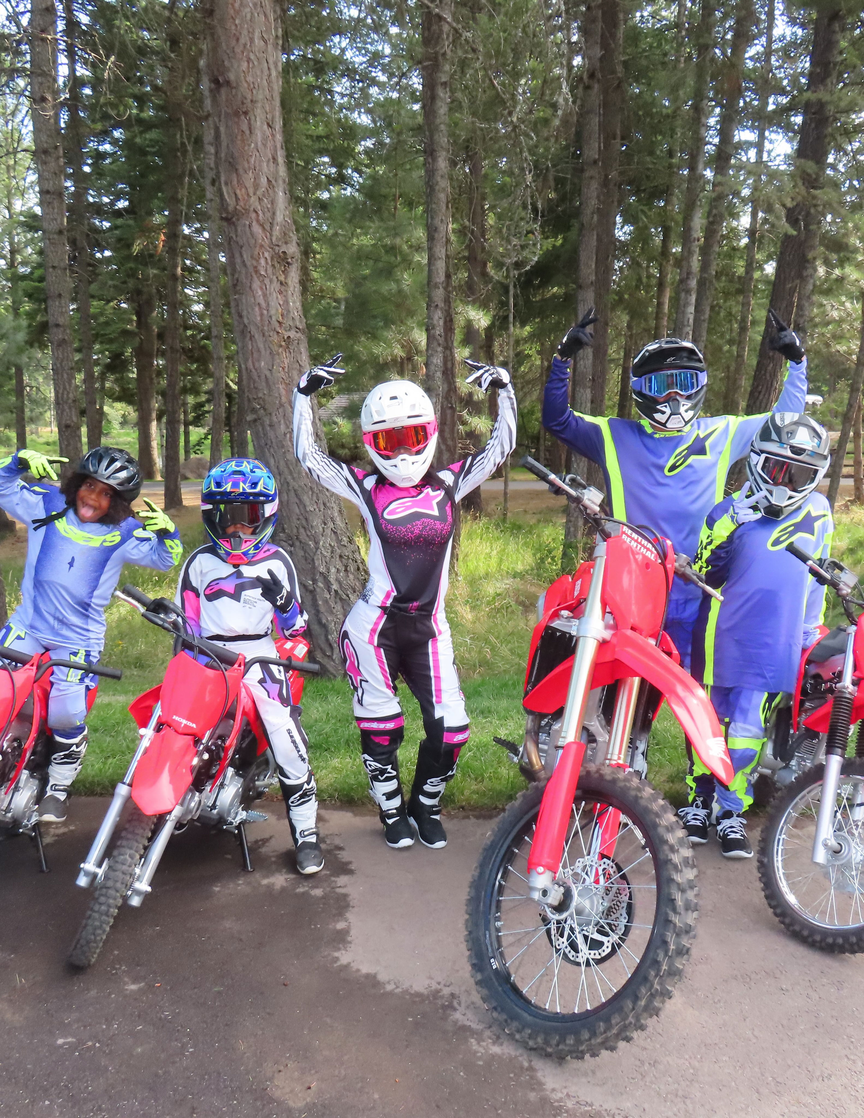 Five people in motocross gear posing with red dirt bikes on a paved surface surrounded by trees.