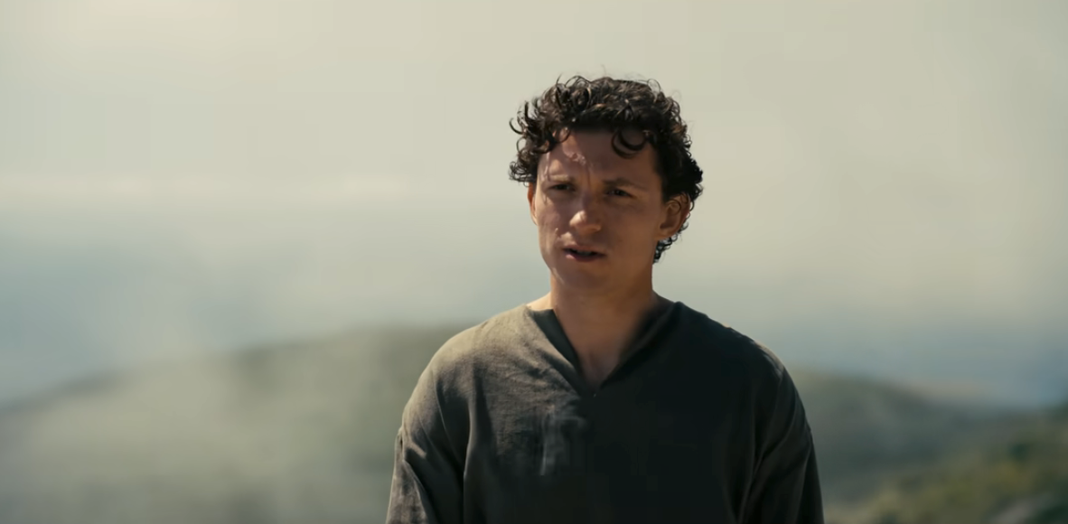 A person stands outdoors with a neutral expression, wearing a casual V-neck shirt, with blurred mountains in the background