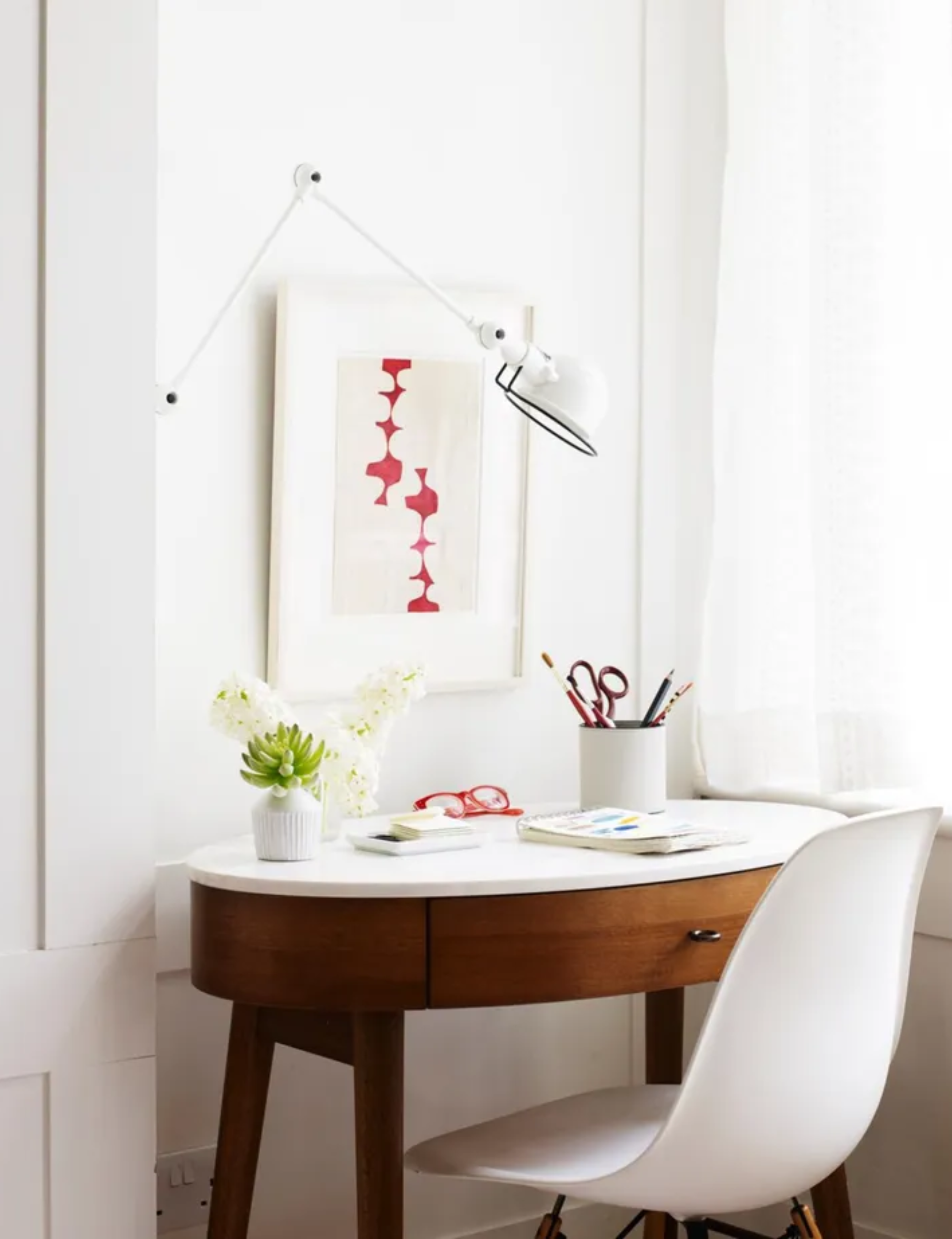 a desk with a white chair, a piece of art hanging on the wall, a wall-mounted lamp, and a cup of pencils on the desk