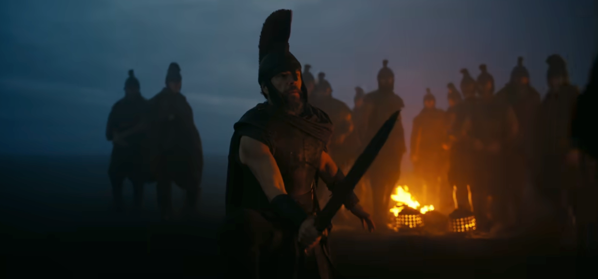 A warrior stands wielding a sword in a dramatic, foggy scene with a campfire and soldiers in the background