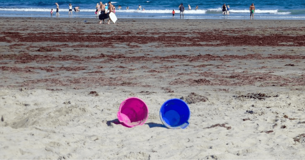 A pink and a blue bucket sit on the sand at Wallis Sands State Park in Rye, New Hampshire as people play in the surf in the background.