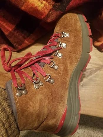 Brown hiking boot with red laces and gray sole, placed on a wooden surface. Cozy wool sock partially visible