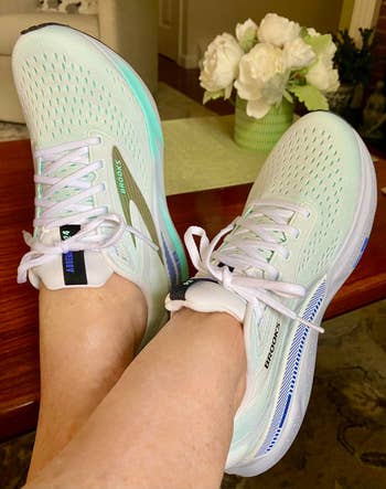 Two feet wearing white Brooks running shoes with mesh patterns, resting on a table. A vase with white flowers is in the background