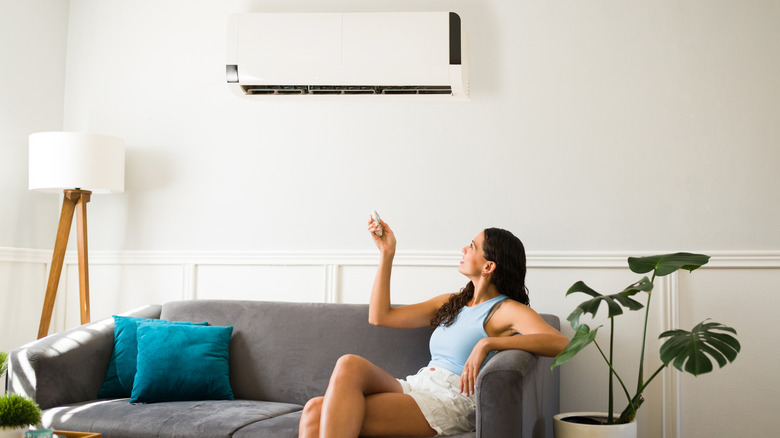 A woman sitting on the couch using a remote to turn on the AC unit above her.